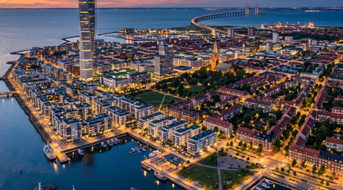 Aerial view of Malmö city with lit buildings, harbor, and the Öresund Bridge at dusk